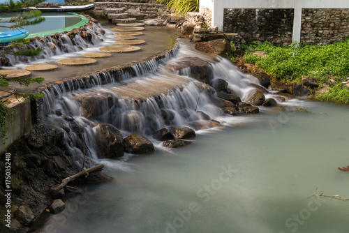 Water flows down over stone steps into a turquoise pond at De'camp hill ka-ang, Nakhon Nayok, Thailand. A tranquil scene of artificial waterfall in tropical-style garden resort.