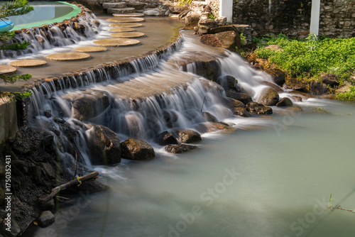 Water flows down over stone steps into a turquoise pond at De'camp hill ka-ang, Nakhon Nayok, Thailand. A tranquil scene of artificial waterfall in tropical-style garden resort.