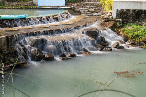 A serene waterfall flows over stone steps with circular stepping stones and paddle boards in a relaxing natural resort setting at De'Camp Hill Ka-ang, Nakhon Nayok, Thailand.