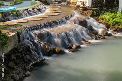 Water flows down over stone steps into a turquoise pond at De'camp hill ka-ang, Nakhon Nayok, Thailand. A tranquil scene of artificial waterfall in tropical-style garden resort.
