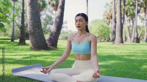 Asian woman meditating in a peaceful park setting, sitting cross-legged on green grass with trees in the background on a sunny day. Concept of mindfulness, self-care, and outdoor relaxation