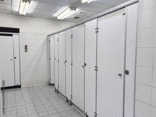 A simple, clean image of a public restroom showing a row of white stalls with metal hardware, a single urinal, and white tiled walls.