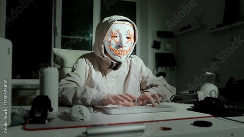 Masked hacker in hood sits at computer, coding at night with glowing mask, concept of cybercrime, online hacking danger, data theft and cyber security breach.