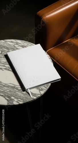 A white book rests on a marble table next to a brown leather chair, casting shadows.