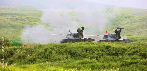 Powerful armored systems demonstrate their formidable firing capabilities on a practice range, creating dynamic smoke trails against the vibrant, expansive natural backdrop of rolling grassy terrain.
