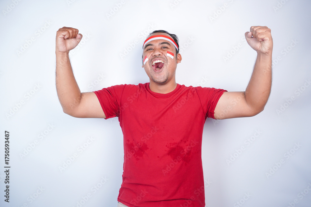 Naklejka premium A young Asian man with a happy successful expression, raised a fist wearing red shirt and flag headband isolated by white background. Indonesia's independence day concept.