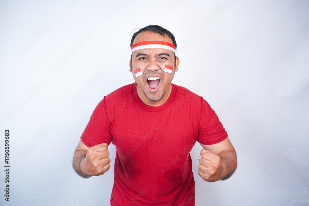 Naklejka premium A young Asian man with a happy successful expression, raised a fist wearing red shirt and flag headband isolated by white background. Indonesia's independence day concept.