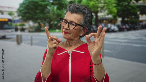 Fotografie Senior woman with grey hair in red coat gesturing ok and pointing while standing outdoors on a city street with a blurred bus in the background