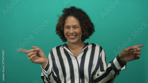 Fotografie Middle aged hispanic woman shrugging shoulders and raising hands in a teal studio wearing a striped blouse and smiling broadly; joy