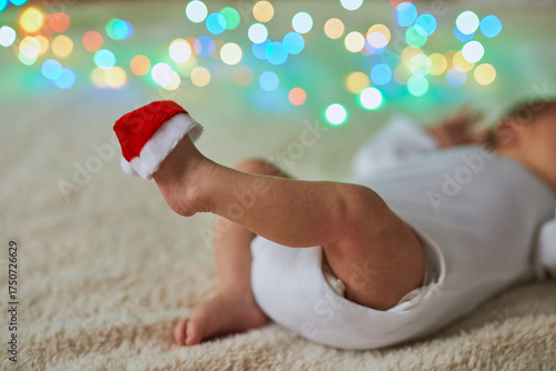 Close-up shot of a newborn baby lying on its back with its tiny feet in the air, one of which has a miniature red Santa hat perched on the toes.