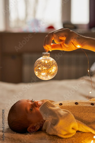 Newborn baby in a knit romper lies on a cozy blanket, gazing up in wonder at a clear Christmas ornament held above it that is glowing with warm fairy lights