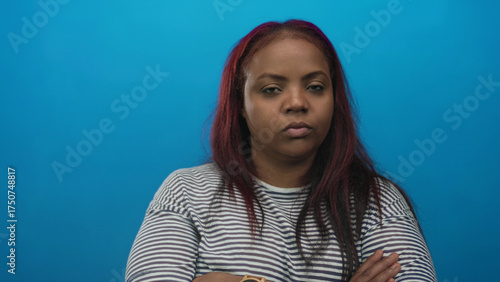 Fotografie Black woman with arms crossed in striped shirt looks unimpressed with neutral gaze in blue studio setting; rejection