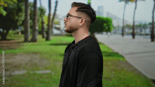 Young man with beard and glasses standing in outdoor park setting surrounded by trees symbolizing relaxation and natural environment in alignment with nature