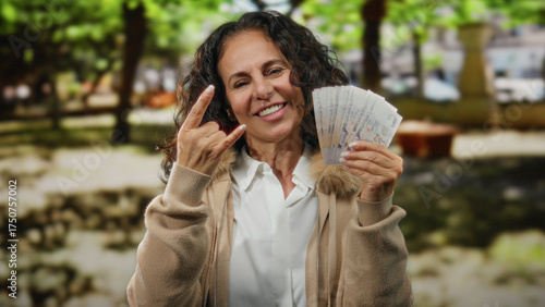 Middle-aged woman holding singapore banknotes making a rock gesture in a sunny outdoor park setting, exuding confidence and joy.