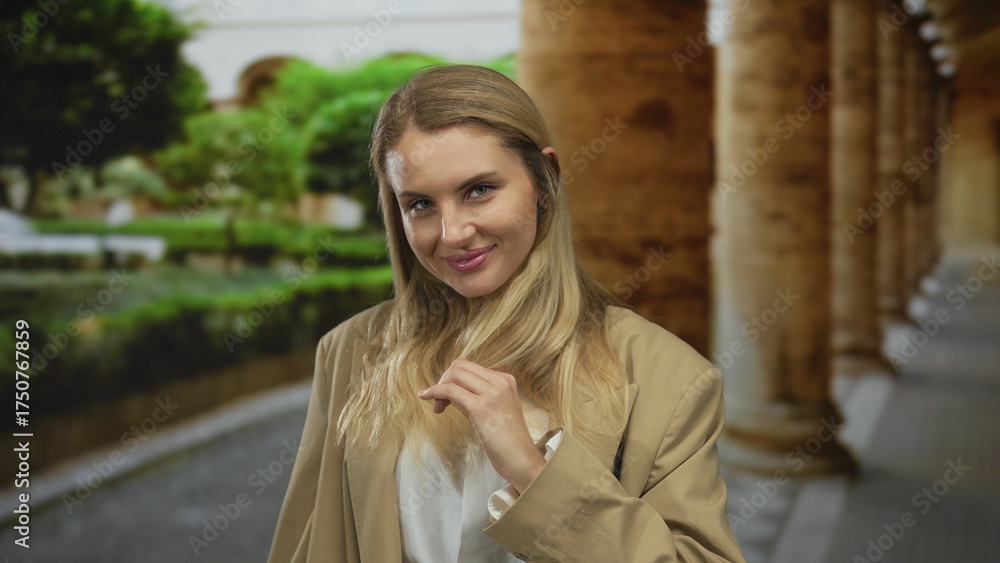 Fototapeta premium Woman smiling outdoors in a lush garden setting with arched architectural elements around, wearing a beige blazer and exuding confidence and warmth.