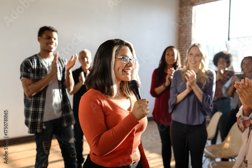 Group applauding a woman speaking with a microphone. Diverse audience clapping. Woman in focus, smiling, engaging with the crowd. Bright, positive atmosphere. Woman presenting in microphone at event.