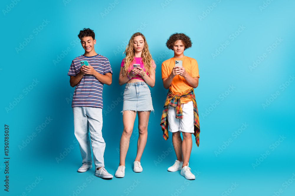 Obraz premium Three teens on a bright blue studio background texting on smartphones smiling and posing as friends showing casual fashion style