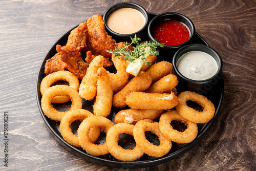фотографія Assorted fried appetizers on a black plate with dipping sauces, including onion rings, mozzarella sticks, and chicken tenders, served on rustic wood background