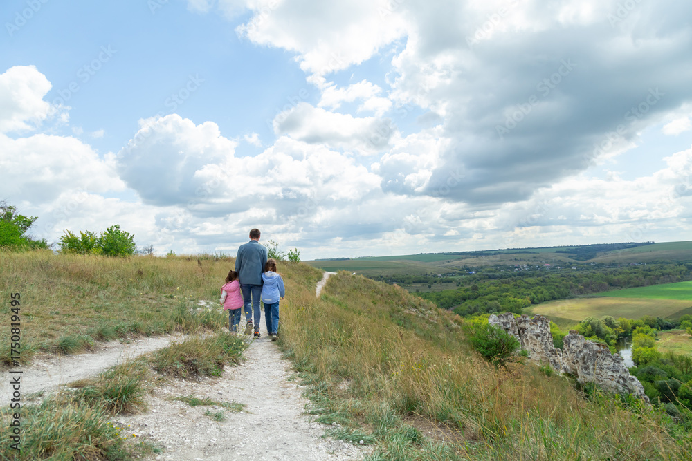 Fototapeta premium Father and daughter stand on top of hill and enjoy beautiful scenery of green hills under blue cloudy sky.