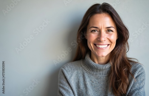 Happy middle aged woman smiles wears gray sweater against gray background. Attractive female with long brown hair looks at camera showing teeth. Portrait of cheerful person.