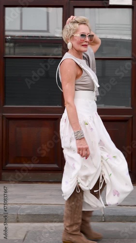 Stylish senior woman in fashionable street outfit walking in city, wearing modern grey top, white skirt, sunglasses and suede cowboy boots, elegant mature fashion model lifestyle portrait