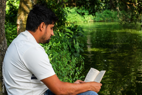 Peaceful moment as a young man enjoys reading a book by the serene lake, surrounded by lush greenery, embracing quiet reflection and intellectual stimulation in nature's embrace
