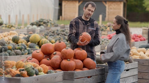 A happy young couple selects a ripe orange pumpkin from a wooden crate at an outdoor farmers market. Seasonal harvest shopping for Halloween or Thanksgiving holiday celebration and decoration.