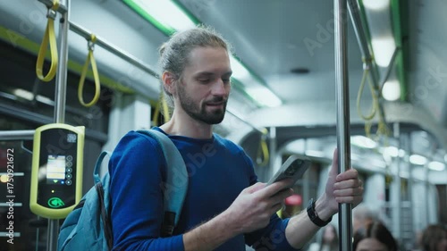 Caucasian man with beard wearing blue sweater standing on city bus holding pole with one hand and smartphone in other. Male reading online news during evening commute home from work.