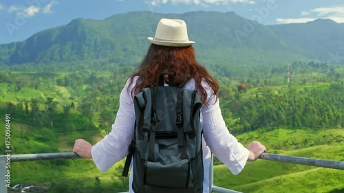 Female traveler standing on green valley landscape, wearing backpack and sunhat, overlooking scenic mountain terrain with lush vegetation and expansive sky
