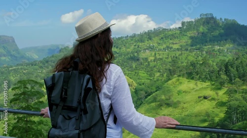 Female tourist with backpack and hat admires the landscape of tea plantations and green hills from a panoramic viewpoint in Ella, Sri Lanka