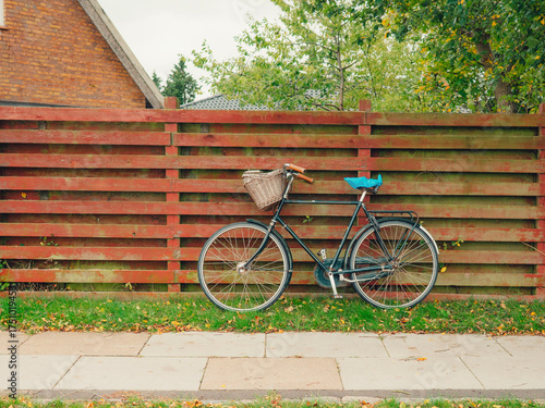 Vintage bicycle with basket rests against a rustic red wooden fence and house in Kastrup, Denmark, embodying Nordic travel and leisure.