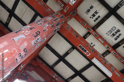 朱塗りの柱と梁に貼られた奉納札のある神社建築。日本の神社建築を見上げる構図。朱塗りの柱と梁に奉納札（千社札）が多数貼られ、歴史と人々の祈りが刻まれたような印象を与える。伝統建築や文化、信仰、時間の積み重ねを感じさせる一枚。