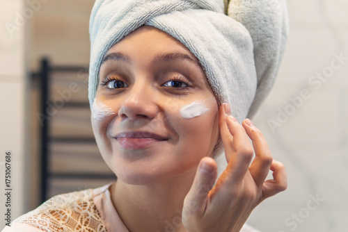 Foto A woman with a towel on her head applies lotion to her face