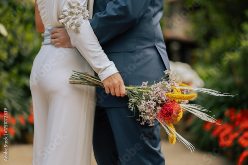 Close-up of bride and groom hugging with hand holding wildflower bouquet, natural style, lush garden background, concept of love and unity