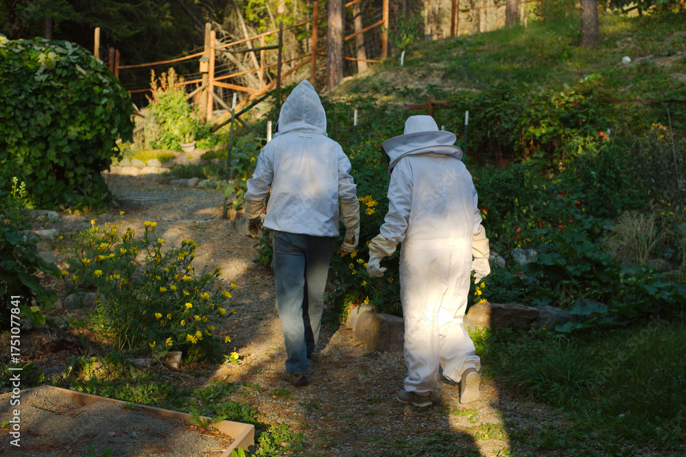 Fototapeta premium Two Beekeepers in full White Protective gear Suits Walk Through a Garden Path Among Flowers. Surrounded by blooming flowers and lush greenery. The scene conveys outdoor work, safety, and harmony with