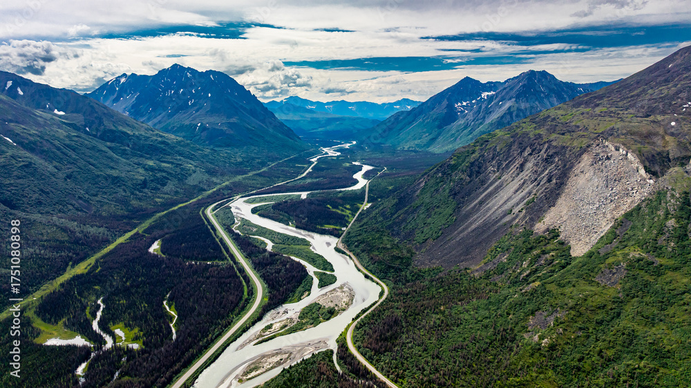 Fototapeta premium Winding river in Alaskan mountain valley aerial view. Aerial view of a winding river between Alaskan mountains with lush green vegetation and cloudy sky.