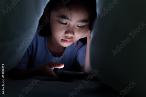 A young Asian girl secretly uses a smartphone under blanket at night. Screen time, addiction, privacy, insomnia, and digital habits among kids concept.