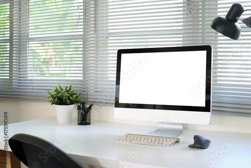 A white computer monitor mockup is placed on a table next to a window in an office.