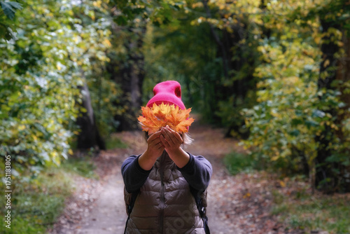 A woman in a red hat and a camisole vest holds a bouquet of autumn leaves, covering her face. Behind her is a blurred path in an autumn park or forest