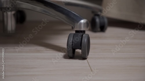 A close-up view captures a dark gray caster wheel, attached to a shiny silver metal chair base, resting firmly on a light-colored wooden floor, highlighting the mobility and stability
