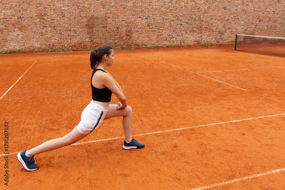 Obraz premium Woman stretching legs on clay tennis court. Concept of sports training, flexibility, preparation and physical wellness through outdoor athletic activity.