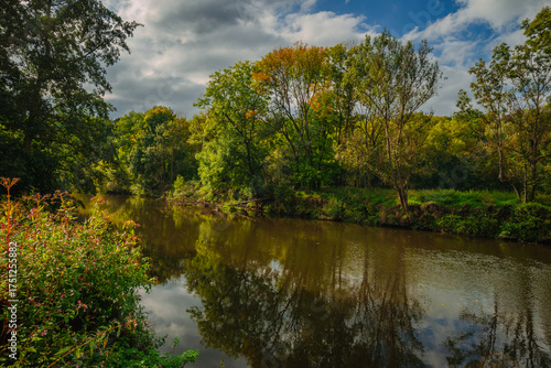 Wide angle view of a slow flowing river surrounded by dense forest with autumn colors under a moody sky where dark clouds contrast with bright sunlight hitting the yellow and green tree canopy.