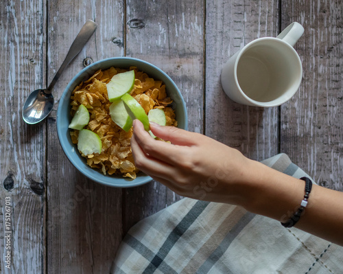 Manipulacion de alimentos, manejo de comida en la cocina, manos tomando trozos de manzana verde para ponerlos en un tazon con cereal, con una taza blanca vacia y una cuchara al lado, vista aerea