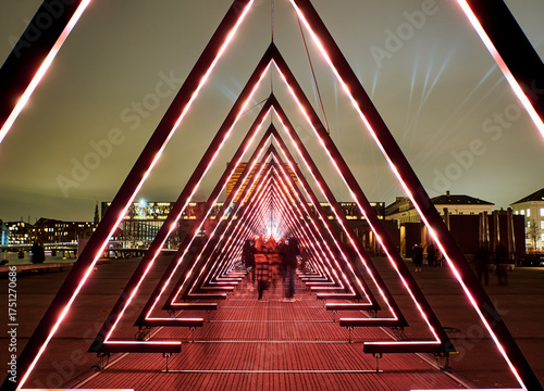 Red led triangles form a striking geometric tunnel at night, with silhouetted figures walking through. The fusion of technology and human presence conveys how modern design transforms public space