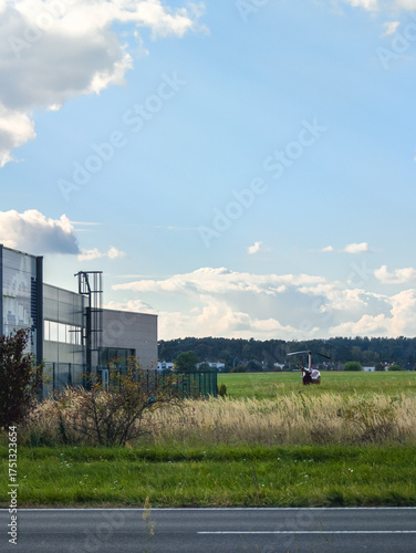 View from a roadside towards a small airfield with a red helicopter parked on the green grass near a modern industrial building under a vast sky with scattered clouds.
