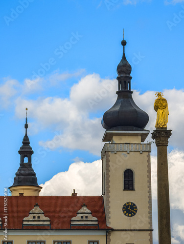 Close-up of a majestic church tower with a black onion dome and a golden statue on a tall column under a blue sky with white clouds and red tiled roofs in the foreground.