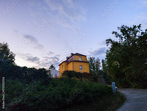 Small bright yellow house standing on a hill surrounded by dense green forest under a pale evening sky in late summer or early autumn.