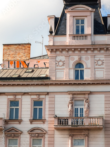 Exquisite architectural details of a pastel pink neo-renaissance building featuring ornate statues on a balcony and decorative wreaths under a dark mansard roof with a golden sun glow.