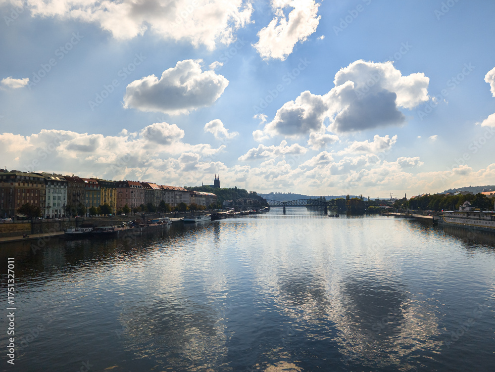 Fototapeta premium Wide angle shot of a calm river under a dramatic cloudy sky with sunlight breaking through reflecting on the water surface between two city shores and a distant metal bridge.