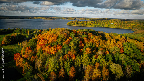 Aerial view of a lake surrounded by a colorful autumn forest, with golden, orange, and green trees glowing under a bright blue sky with scattered clouds.